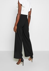 Svart byxdress med breda ben och en vit volanglös topp utan ärmar, med en figurnära midja och dragkedja bak. Svarta högklackade sandaler.