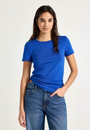 Nuori nainen, jolla on pitkät tummat hiukset, sininen lyhythihainen paita ja siniset farkut, seisoo käsi taskussa yksinkertaisen taustan edessä.