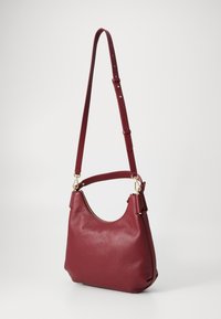 Sac à bandoulière en cuir rouge avec une finition texturée, une forme incurvée, une bandoulière réglable et des accessoires en métal doré, avec une fermeture zippée sur le dessus.