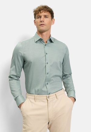 Mann trägt ein hellgrünes Hemd mit Knöpfen und beige Hosen, steht mit den Händen in den Taschen vor einem schlichten hellen Hintergrund.