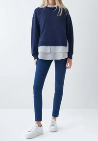 Felpa blu navy indossata sopra una camicia a righe bianche e blu con bottoni, abbinata a jeans skinny blu e sneakers bianche con suola crema.