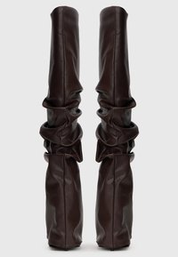 Botas de cuero marrón oscuro de alta textura con un diseño deslavado y punteras afiladas, de pie sobre un fondo liso.