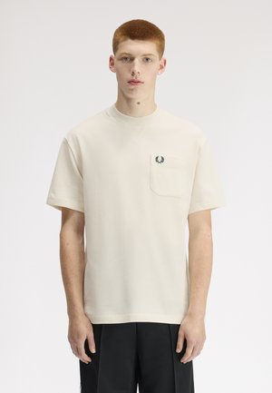 Jeune homme aux cheveux roux courts portant une chemise blanche propre à manches courtes avec un petit logo de couronne noire sur la poche gauche de la poitrine.