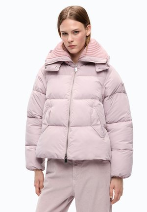 Donna che indossa una giacca imbottita rosa chiaro con un collo alto a coste e pantaloni rosa chiaro abbinati, in piedi davanti a uno sfondo neutro.