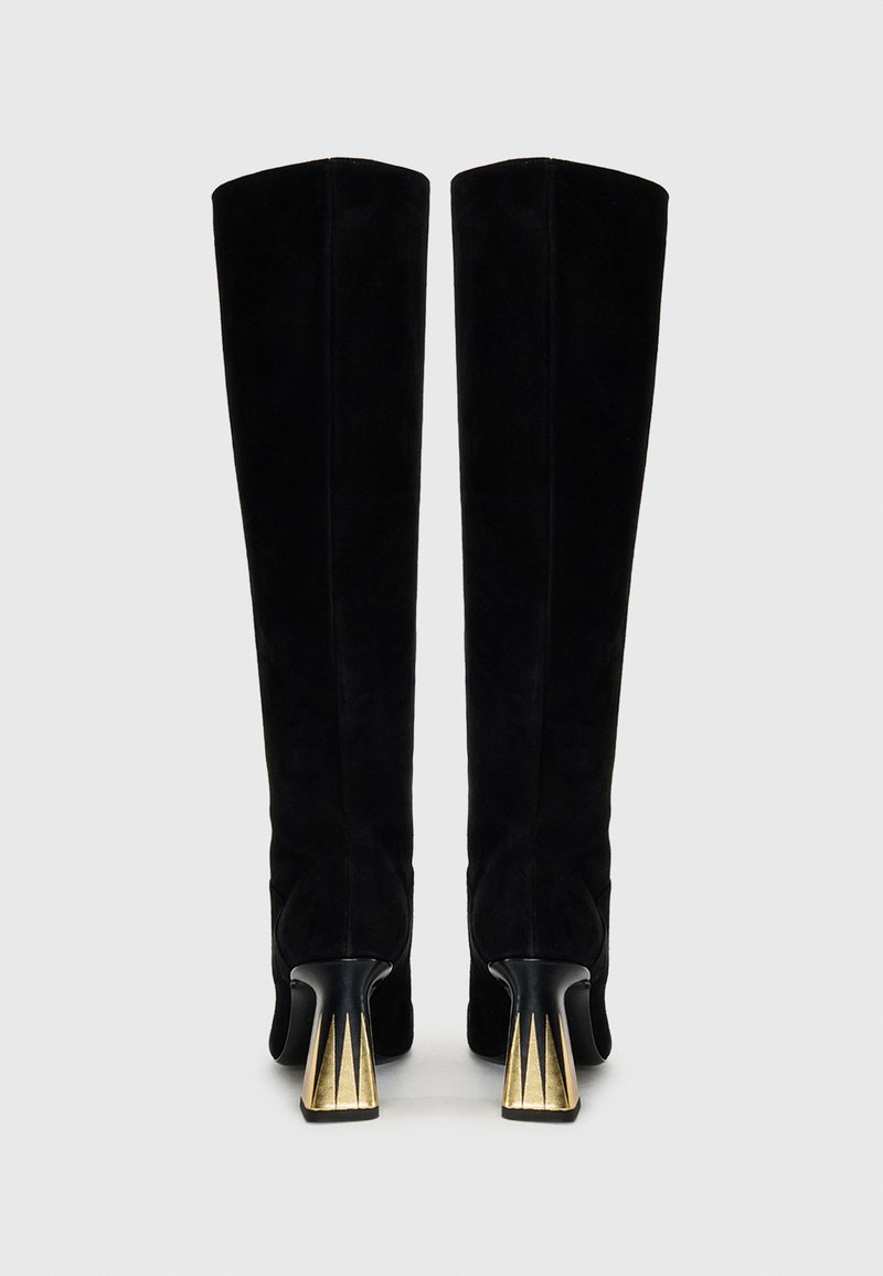 Knöchelhohe schwarze Wildlederstiefel mit glatter Textur und eckigen, goldakzentuierten Absätzen, die ein dreieckiges Muster aufweisen. Klassisches Design.