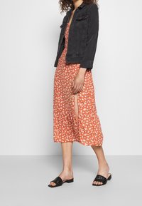 Kvinna i orange blommig midiklänning med sidslits, svart denimjacka och svarta låga vävda sandaler som står mot en neutral bakgrund.