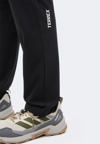 Pantaloni neri con logo "TERREX" sopra scarpe da trail beige, grigie e verdi che hanno anch'esse il marchio "TERREX" sul lato.