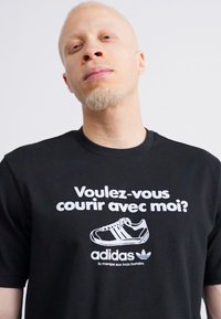 Fekete póló fehér grafikai szöveggel: "Voulez-vous courir avec moi?" és egy fekete futócipő illusztrációval, alatt pedig az Adidas logóval.