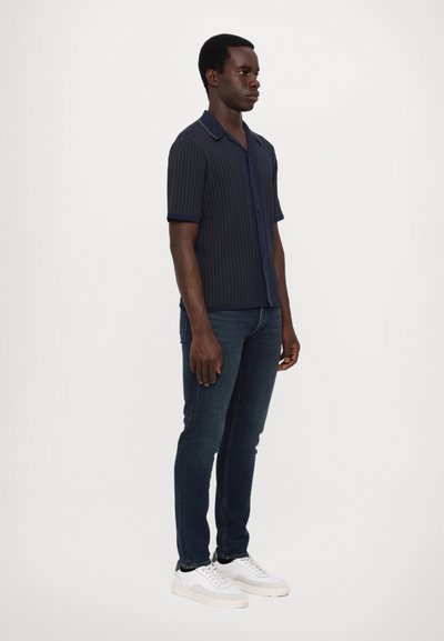 Navy stripete kortermet skjorte med spiss krage, knappefront, kombinert med mørke jeans og hvite joggesko, satt mot en enkel bakgrunn.
