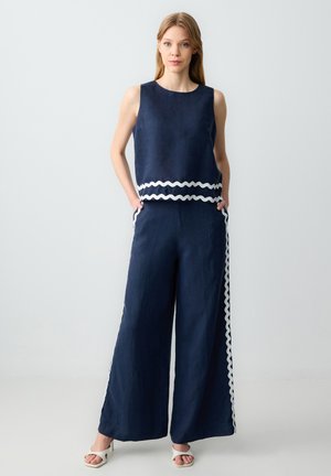 Tuta navy con pantaloni a gamba larga, top senza maniche, bordo ondulato bianco in vita e ai lati, realizzata in tessuto leggero, dotata di tasche laterali.