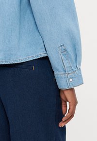 Ανοιχτό μπλε τζάκετ από τζιν με κουμπί στον μανίκι, σε συνδυασμό με σκούρο μπλε υφασμάτινο παντελόνι. Ορατές ραφές τονίζουν το σχέδιο.