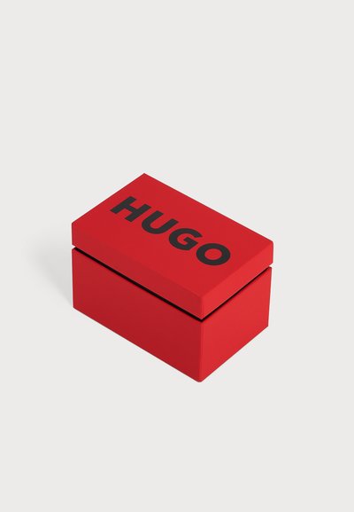 Boîte rectangulaire rouge avec des lettres capitales noires "HUGO" sur le couvercle, placée sur un fond gris clair uni.