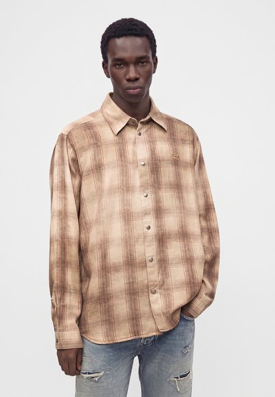 Jeune homme portant une chemise à carreaux beige boutonnée et un jean bleu clair déchiré, debout avec une main dans la poche devant un fond uni.
