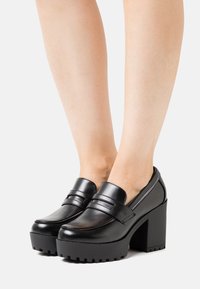 Černé robustní mokasíny na podpatku s platformovými podrážkami, nošené na bosých nohou na jednotném světlém pozadí.