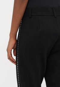 Zwarte broek met een gladde textuur, voorzien van een tailleband en decoratieve stiksels langs de zijkant, die een aansluitende silhouet accentueert.