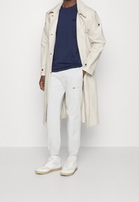 Trench beige, t-shirt blu navy, pantaloni jogger bianchi con logo e sneaker bianche con dettagli beige. Texture lisce e vestibilità sartoriale.