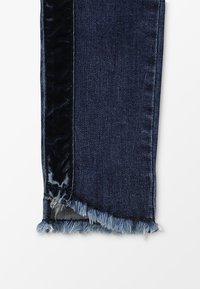 Vaqueros de mezclilla azul oscuro con un panel lateral de terciopelo, dobladillo deshilachado y un contraste de textura distintivo entre los materiales.