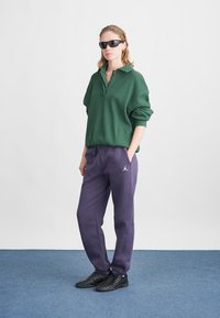 Polo verde texturizado de manga larga con cuello y dos botones, combinado con pantalones de chándal holgados de color púrpura oscuro que presentan un logo. Zapatos negros.