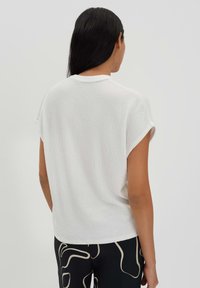 Top corto a maniche corte bianco con un collo rotondo, caratterizzato da una vestibilità rilassata e un tessuto morbido e traspirante. Vista posteriore che mostra un motivo sottile.