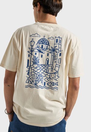 T-shirt en coton couleur crème avec un motif graphique bleu représentant une scène méditerranéenne avec une église, des palmiers et des motifs géométriques.