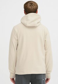 Beige hættebluse lavet af blødt, tekstureret stof, med snoretræk i hætten og ribbede manchetter. Bagfra ses et strømlinet design uden grafik.