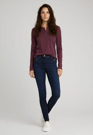 Mujer con cabello largo castaño, vestida con una camiseta de manga larga color granate, jeans azul oscuro y zapatillas blancas, de pie contra un fondo neutro.