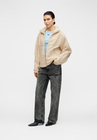 Veste zippée en polaire beige clair avec col montant, associée à un t-shirt bleu et un jean noir large déchiré. Bottes noires à bouts pointus.