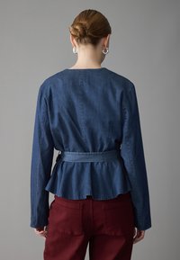 Giacca peplum in denim blu scuro con maniche lunghe e vita legata, abbinata a pantaloni bordeaux a vita alta. Tessuto liscio, senza fantasie.