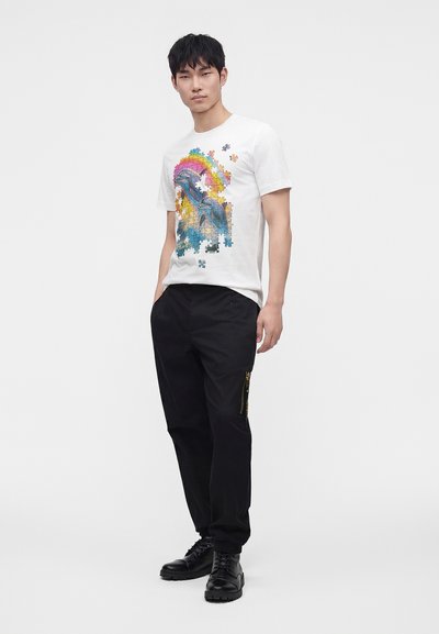 Άνδρας που φοράει λευκό T-shirt με πολύχρωμο σχέδιο πουλιού σε σχήμα παζλ, μαύρο παντελόνι με φερμουάρ και μαύρα δερμάτινα μποτάκια με κορδόνια.