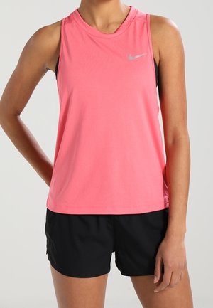 Vrouw draagt een mouwloze roze sporttop en zwarte short, staand met één hand op de heup tegen een effen achtergrond.