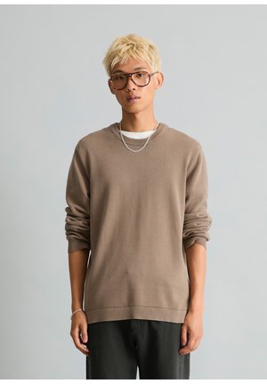 Junge Person mit kurzen blonden Haaren, die große Brille, einen braunen Pullover, schwarze Hose und eine silberne Kettenkette sowie ein Armband trägt, steht vor einem schlichten Hintergrund.