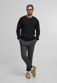 Maglione blu navy a maglia cable-knit, pantaloni grigi a vestibilità rilassata, scarpe beige senza lacci e calze bianche; in piedi con le mani nelle tasche su uno sfondo semplice.