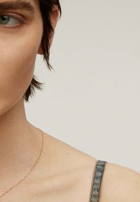 Un primer plano de la clavícula y el hombro de una persona, que presenta una delicada cadena de oro y una correa de denim azul. El fondo es de un color neutro.