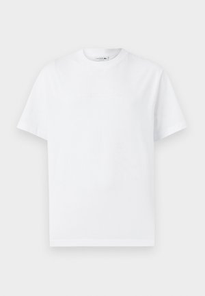 Camiseta de algodón blanca con un clásico cuello redondo, mangas cortas y texto en relieve "Lacoste Heritage" en la parte frontal. Diseño simple.