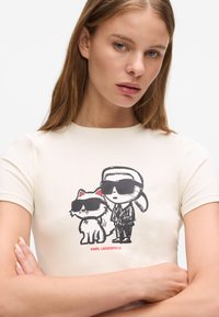 Tricou roșu-crem, ajustat, cu un grafic de desen animat în culorile negru și roșu, reprezentând o pisică și o figură cu ochelari de soare, având inscripția "KARL LAGERFELD."
