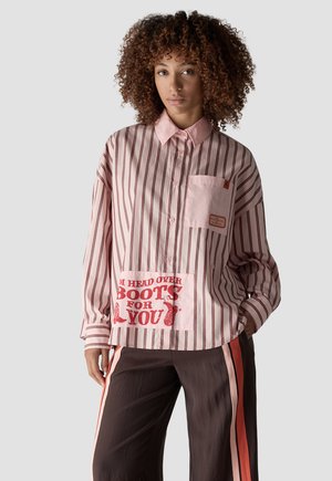 Woman with curly hair wearing a pink striped shirt with red text patch and dark pants with pink and red side stripes against a plain background.