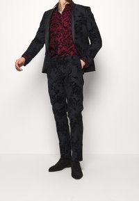 Musta, kukkakuvioitu puku, joka on valmistettu teksturoidusta kankaasta, yhdistetty viininpunaiseen paitaan, jossa on samanlaisia design-elementtejä. Asun täydentävät mustat kengät.