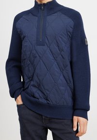 Pull bleu marine à demi-fermeture éclair, avec un devant matelassé et des manches ainsi qu'un bas en tricot côtes. Motif en losanges remarquables sur le tissu.