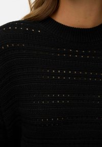 Maglione lavorato a maglia nera con strisce orizzontali e piccoli fori aperti, scollatura a coste e tessuto textured. Inquadratura ravvicinata del materiale.