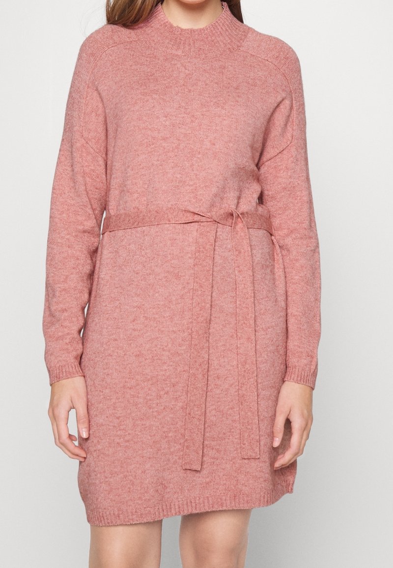 Femme portant une robe en maille rose à manches longues, longueur genou, avec un col montant et une ceinture nouée à la taille.