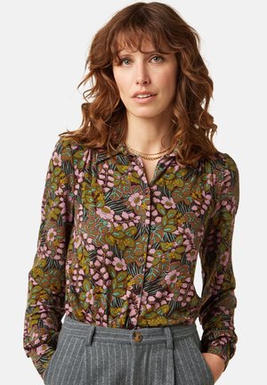 Vrouw met krullend roodbruin haar, die een gebloemd hemd met knopen draagt in groene, roze en bruine tinten en een grijze broek met fijne krijtstrepen.