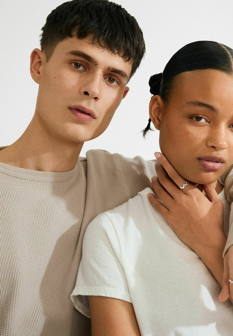 In primo piano, un uomo e una donna si avvicinano; l'uomo indossa una camicia beige con una texture, mentre la donna ha una camicia bianca e tiene una mano sul collo, indossando anelli ondulati.