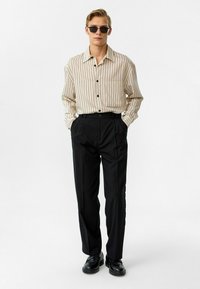 Gestreepte, beige knopoverhemd met een borstzak, gecombineerd met zwarte geplooide broeken en zwarte schoenen, gedragen met een zonnebril.