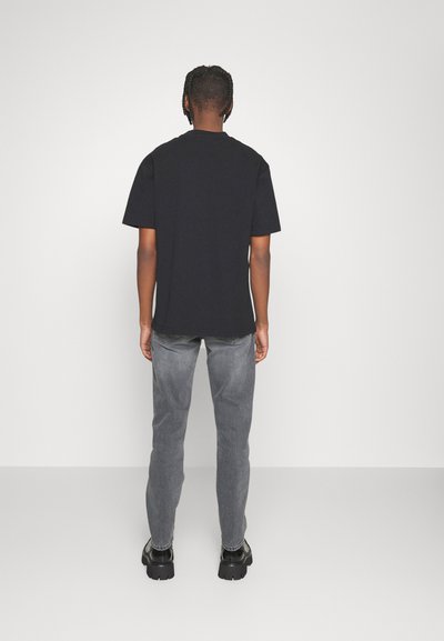 T-shirt de algodão preta com mangas curtas, vista de costas, combinada com jeans de ganga cinza e sapatos pretos de sola grossa.