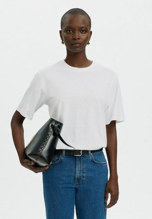 Vrouw met wit T-shirt en blauwe jeans houdt een zwarte leren handtas met veterdetail vast tegen een effen achtergrond.