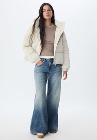 Veste matelassée de couleur crème avec capuche, associée à un jean évasé bleu clair. Le tissu semble lisse et légèrement brillant.