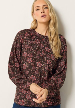 Vrouw met lang blond haar die een donker gebloemde blouse draagt met roze bloemen en lange mouwen, staand tegen een effen lichte muur.