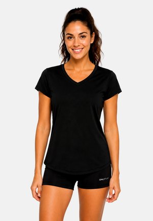 Frau mit dunklen Haaren im Pferdeschwanz, trägt ein schwarzes V-Ausschnitt-T-Shirt und schwarze Shorts, lächelnd und vor einem einfarbigen Hintergrund stehend.