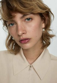 Camicia beige con colletto e dettaglio con zip, indossata da una modella con capelli corti e ondulati e orecchini a cerchio argentati. Sfondo neutro.