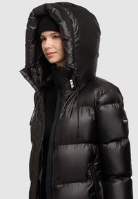 Melna pūtēja jaka ar lielu kapuci, spīdīga tekstūra, priekšējā rāvējslēdzēja un regulējamas auklas. Kreisajā piedurknē ir logotipa uzlīme.